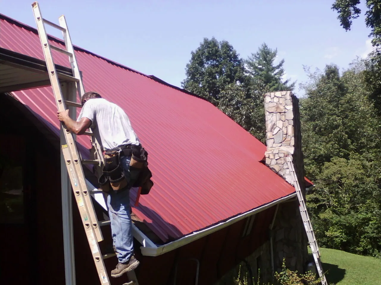 Roofer on a ladder beside a red metal roof and stone chimney — Soffit & Fascia Repair by Roofing Brothers in Beemer